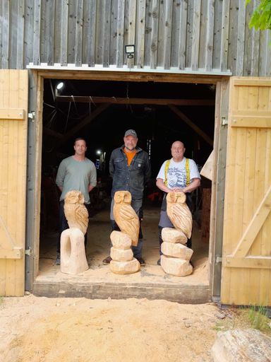Drei Männer stehen vor einer Holzscheune im Erzgebirge, jeder neben einer selbst geschnitzten Holzeule aus dem Kettensägen-Schnitzkurs von A