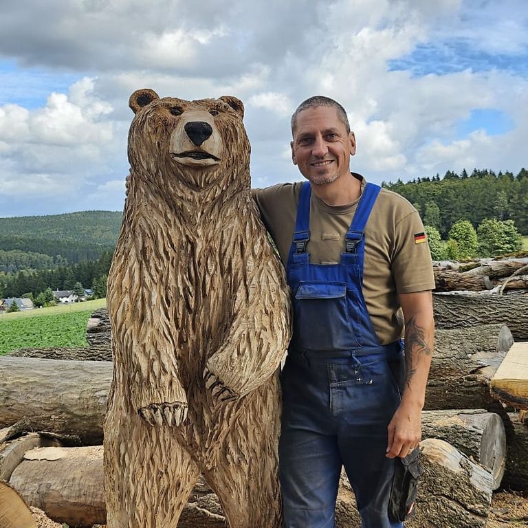 Ein Kettensägenkünstler in Arbeitskleidung steht neben einer detailreich geschnitzten Holzskulptur eines Bären im Freien, umgeben von Baumst