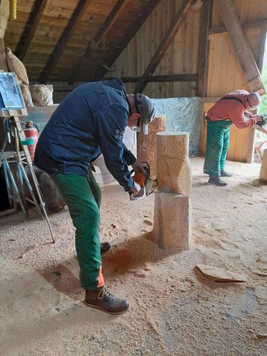 Kursteilnehmer in orangener Schnittschutzhose schnitzt mit einer Husqvarna-Kettensäge an einem Holzstamm.